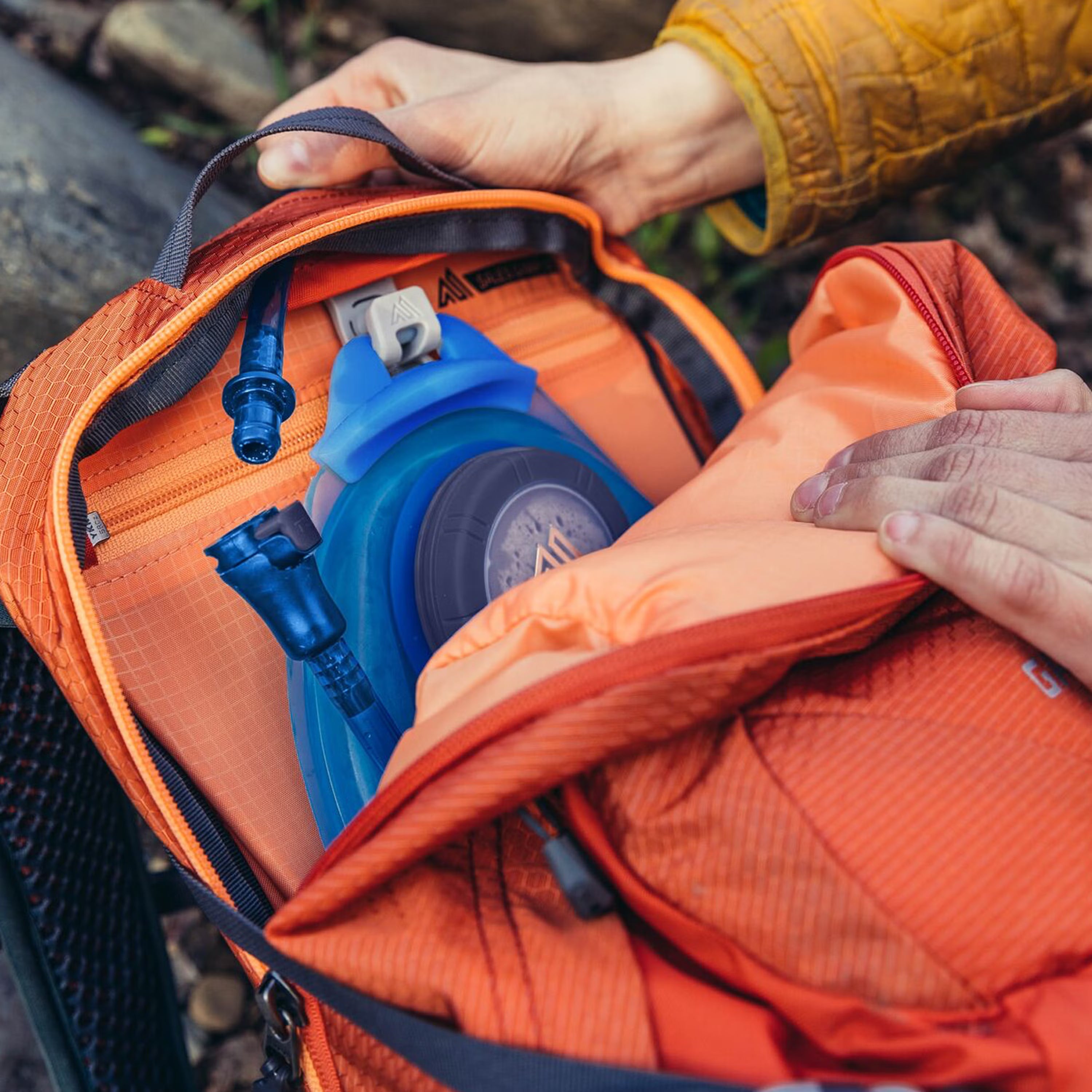 Person opening an orange backpack with a blue water bottle inside, outdoors.